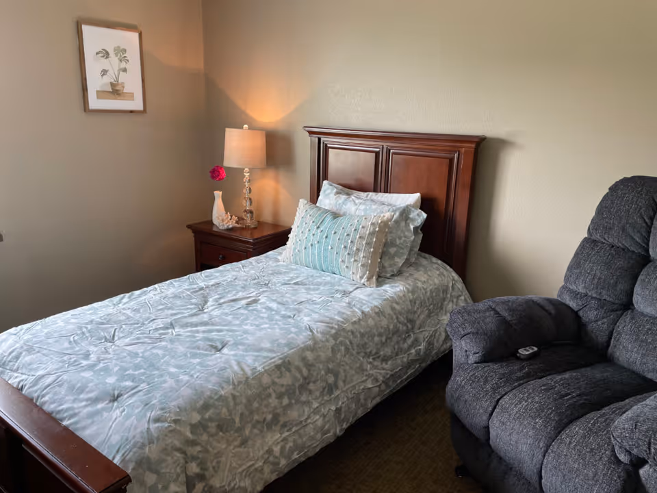 A cozy bedroom with a single wooden bed featuring light blue patterned bedding and multiple pillows. Next to the bed is a wooden nightstand with a decorative lamp, a white vase with a pink flower, and a small coral decoration. A framed botanical print hangs on the beige wall above the nightstand. To the right of the bed is a dark gray recliner chair with a remote control on the armrest.