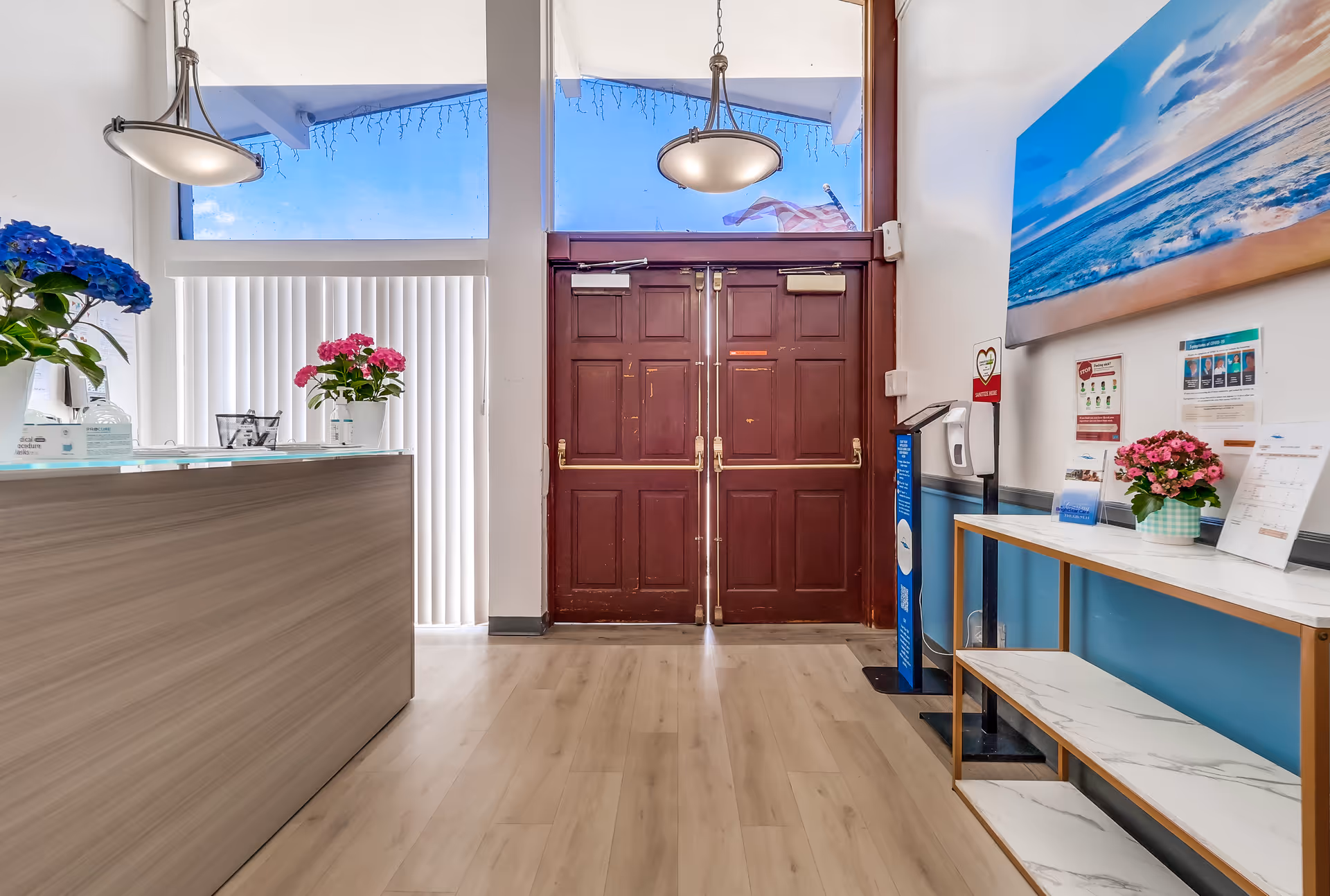 Interior view of a facility entrance with double wooden doors, a reception desk on the left with a blue flower pot, and a white and gold shelf on the right with a pink flower pot and informational pamphlets. Two hanging ceiling lights are visible, and a large ocean-themed painting is mounted on the right wall.