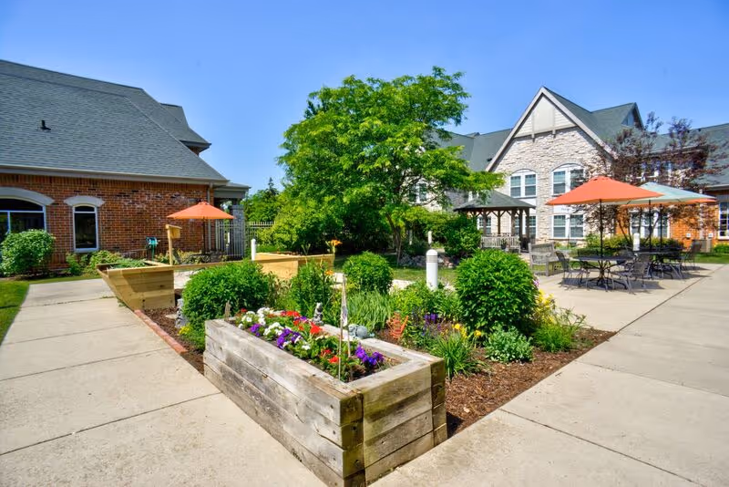 Outdoor garden area at Newcastle Place featuring raised wooden flower beds with colorful flowers, green shrubs, and a large tree. There are paved walkways, patio tables with orange umbrellas, and brick and stone buildings in the background under a clear blue sky.