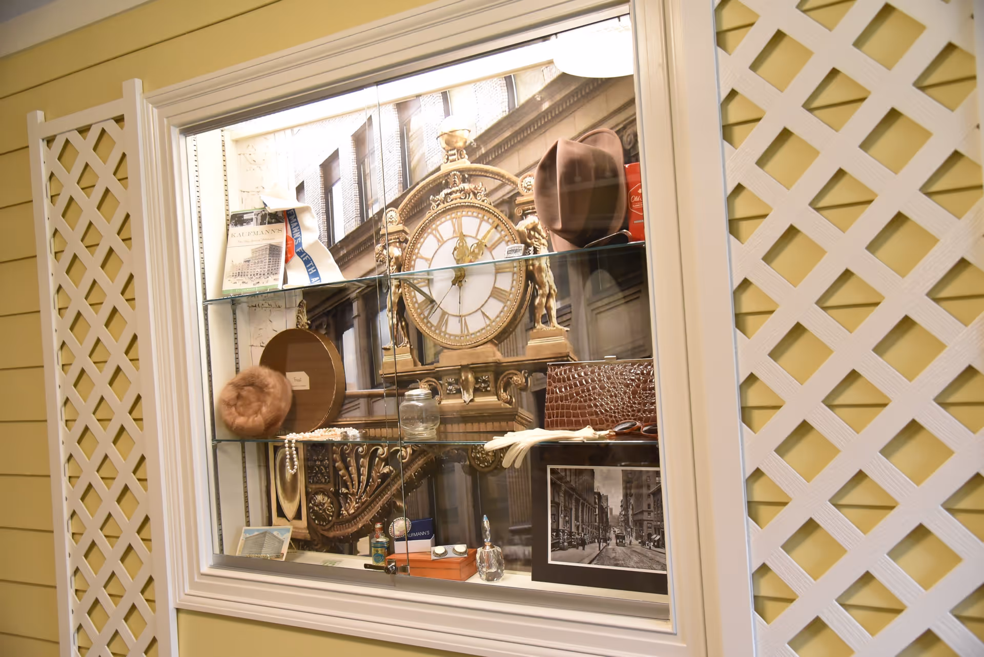 A glass display case mounted on a yellow wall with white lattice panels on either side. Inside the case are various vintage items including a brown hat, a pearl necklace, a framed black and white photograph of a street scene, a small glass jar, a brown textured handbag, white gloves, and a large ornate clock face in the background.