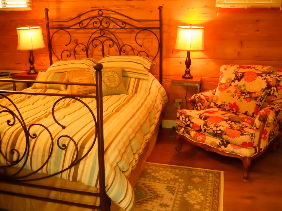 Cozy bedroom with a wrought-iron bed, striped bedding, two bedside lamps and a floral armchair against wood-paneled walls.