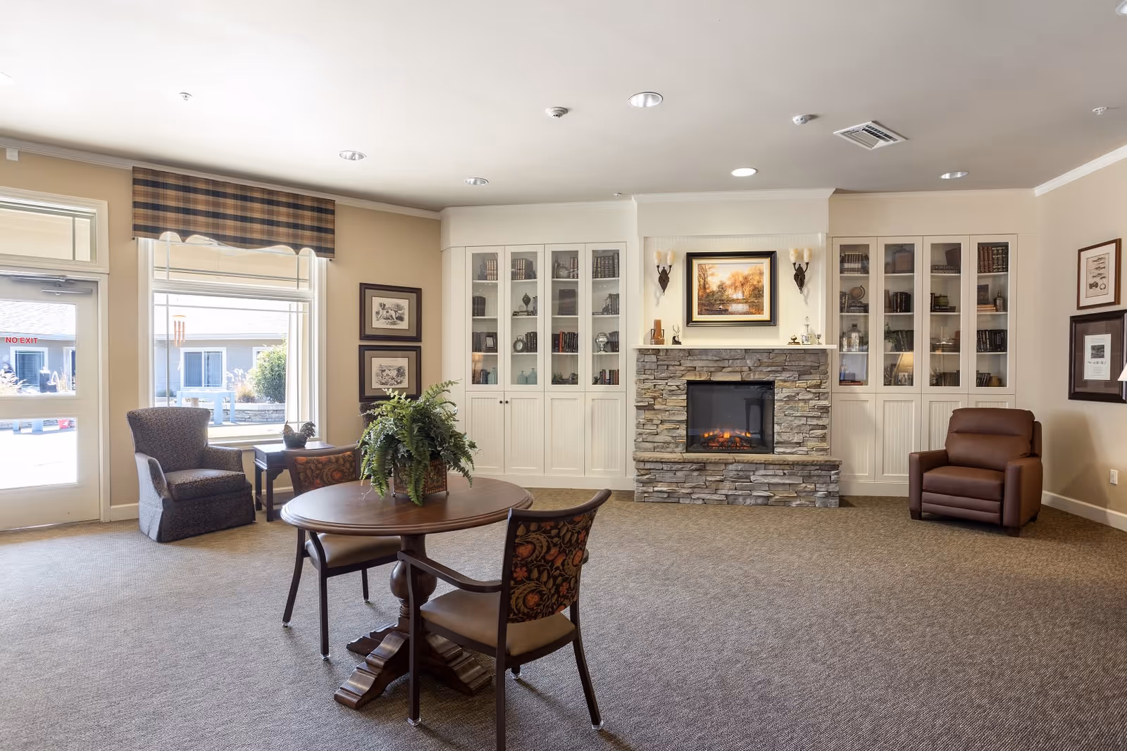 A cozy living room area in an assisted living facility featuring a stone fireplace with a painting above it, built-in white cabinets with glass doors displaying books and decorative items, a round wooden table with two chairs, a potted plant centerpiece, a comfortable armchair near a window with plaid valance, and another brown recliner chair against the wall.