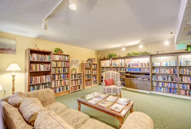 A cozy library room with multiple wooden bookshelves filled with books along the walls. There is a patterned armchair with a red pillow and a wooden coffee table in the center with magazines or books on it. A beige couch is partially visible in the foreground. The room is softly lit with ceiling lights and a floor lamp, and there are some decorative plants on top of the bookshelves.