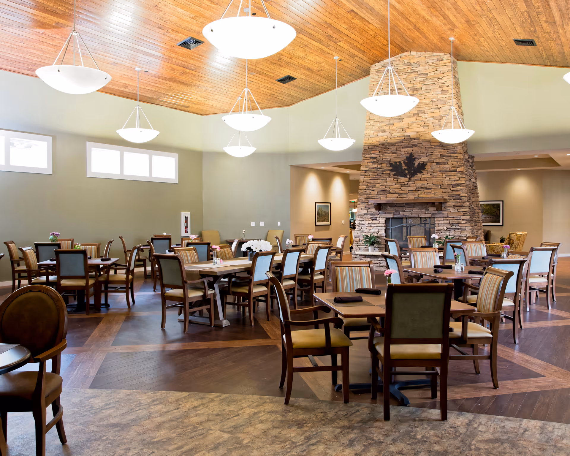 Spacious dining room with multiple tables and chairs, a stone fireplace, and pendant lights under a wood-paneled ceiling.