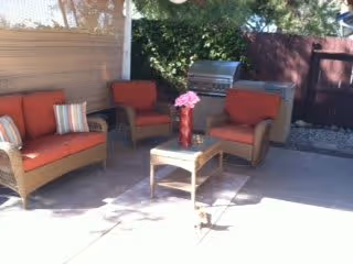 Outdoor patio area with a set of wicker furniture including a loveseat, two armchairs with red cushions, and a small table with a vase of pink flowers. A barbecue grill is visible in the background along with trees and a fence.