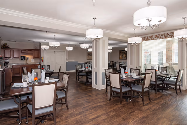 A spacious dining room in an assisted living community featuring multiple wooden tables with chairs arranged around them. Each table is set with white napkins, cups, and small floral centerpieces. The room has wooden flooring, soft lighting from ceiling chandeliers, large windows with blinds, and a kitchen area with cabinets in the background.