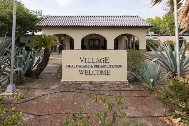 Front exterior view of Village Healthcare & Rehabilitation building with a welcome sign in front, surrounded by plants and trees under a partly cloudy sky.