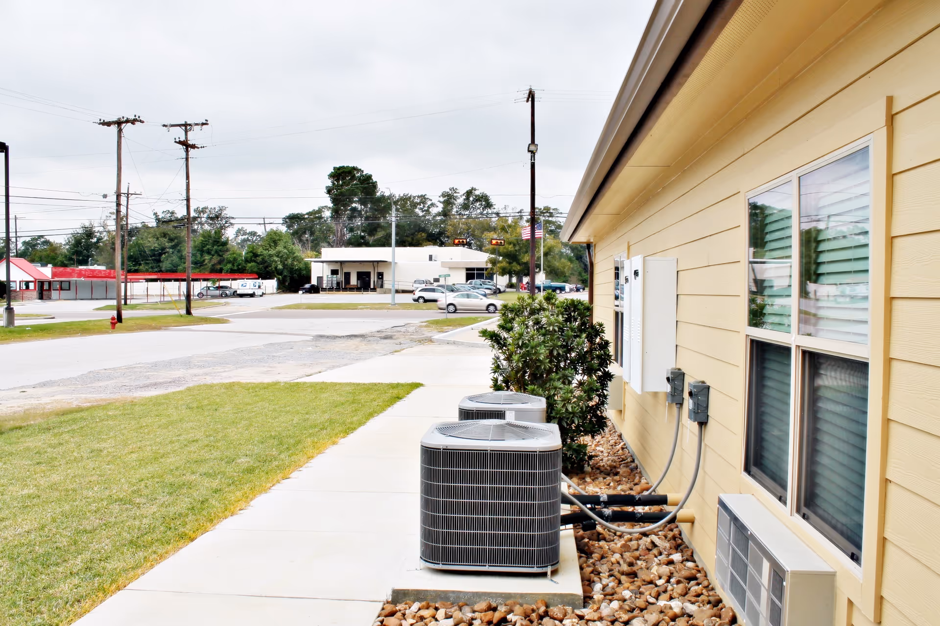Outdoor view alongside a yellow building showing two air conditioning units on a bed of rocks next to a sidewalk. There is a small bush near the units and a window with blinds on the building. In the background, there is a street with cars, utility poles, and other buildings under a cloudy sky.