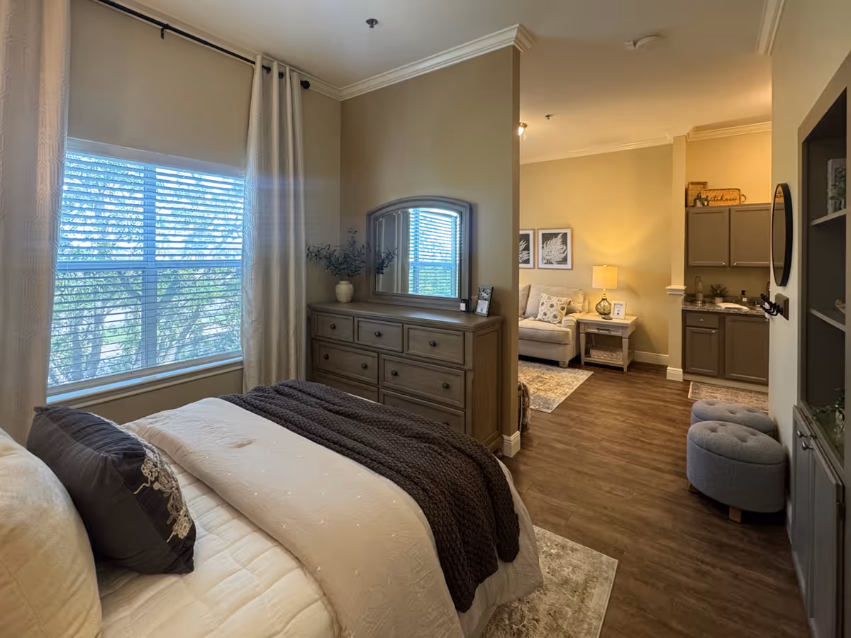 A cozy bedroom with a large window covered by curtains and blinds, a bed with pillows and a dark throw blanket, and a wooden dresser with a mirror and decorative plant. The room opens into a living area with a sofa, side table with a lamp, and a small kitchenette with cabinets and a countertop.