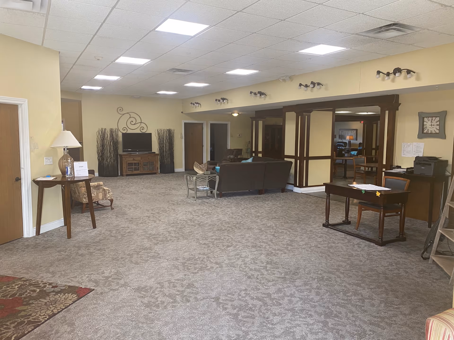 A spacious common area in a senior living facility with beige walls and patterned carpet. The room features a brown couch, a glass coffee table, a TV on a wooden stand flanked by decorative tall branches, a small wooden desk with a chair, and a table with a lamp and hand sanitizer. There are multiple ceiling lights and wall sconces providing illumination. A large mirror with wooden framing is on one wall, and a clock is mounted above a printer on a side table.