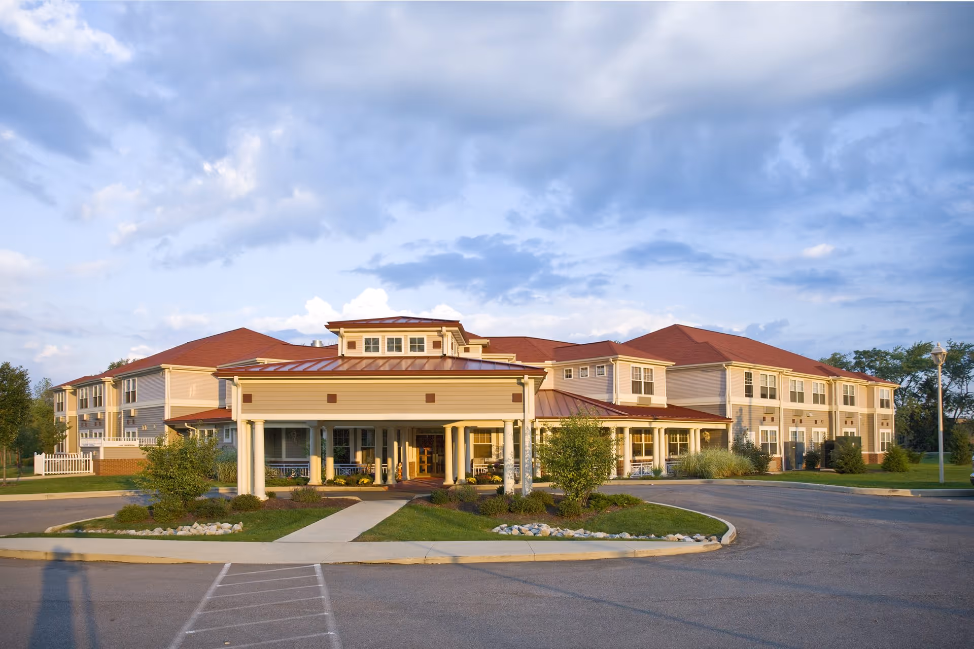 Exterior front view of a large senior living facility building with beige siding and red roofs under a partly cloudy sky. The entrance has a covered driveway with white columns and landscaped greenery around the walkway.