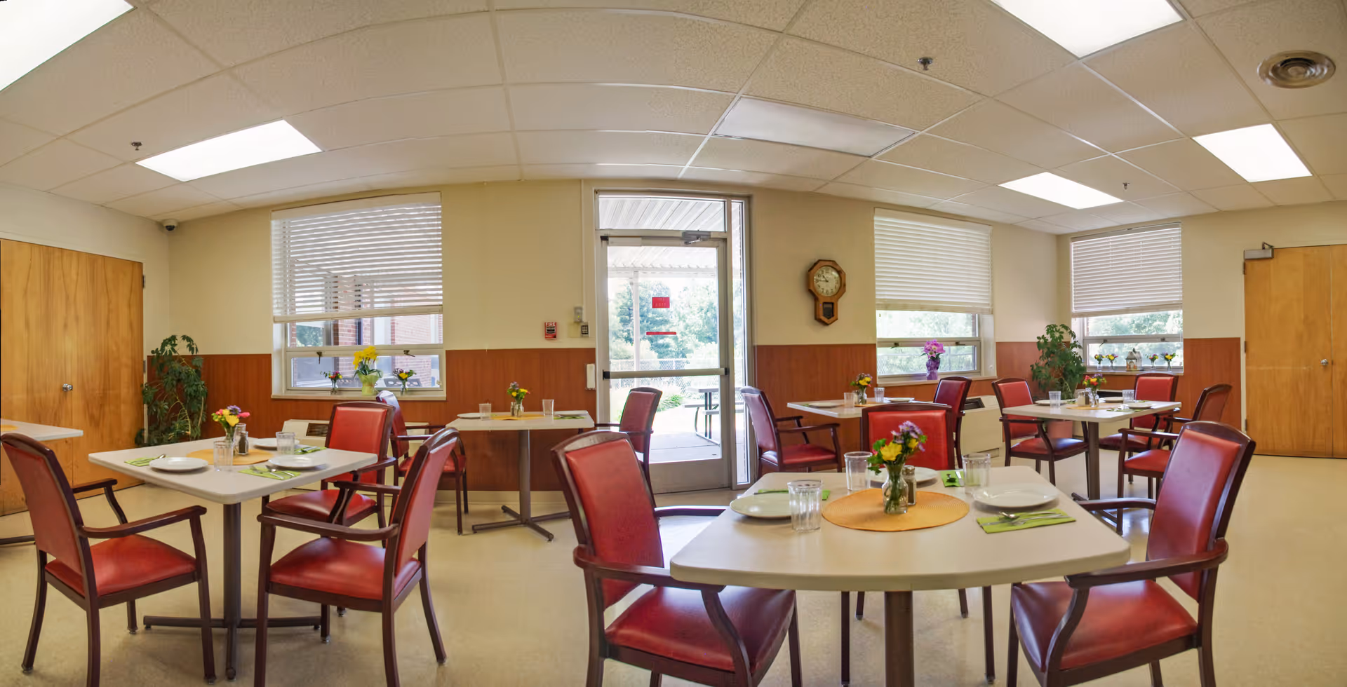 A bright dining room with several square tables set with plates, glasses, and small flower vases. The room has red cushioned chairs, large windows with blinds, a wooden clock on the wall, and a door leading outside. There are plants placed near the windows and doors.