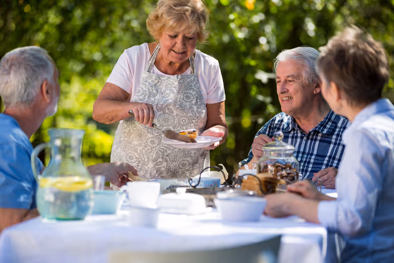 A group of elderly people sitting around a table outdoors, with one woman standing and serving a slice of pie to a man. The table is set with cups, a pitcher of water with lemon, and a jar of cookies, surrounded by greenery in the background.