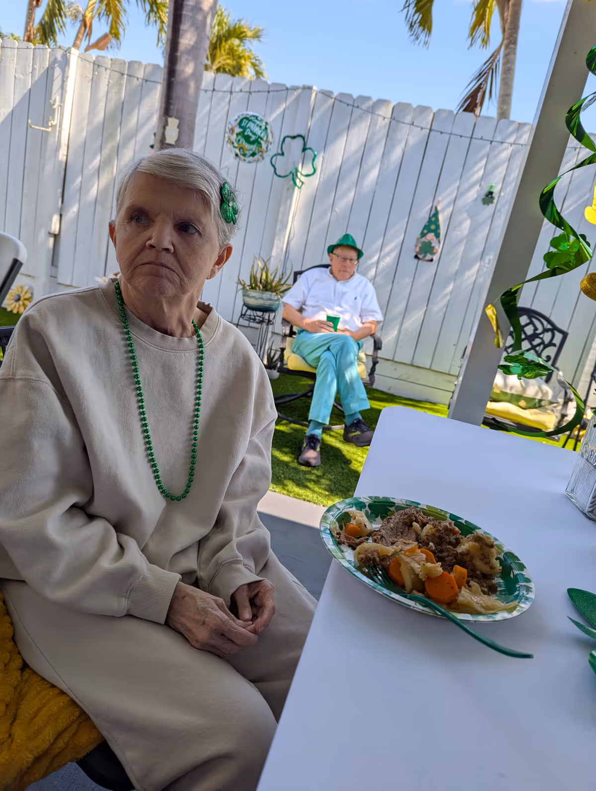 An elderly woman wearing a beige sweatshirt and green beaded necklace sits at a table with a plate of food in front of her. In the background, an elderly man wearing a green hat and light blue pants sits on a chair near a white wooden fence decorated with St. Patrick's Day decorations including shamrocks and a sign. The setting appears to be an outdoor patio area with green artificial grass and palm trees visible.