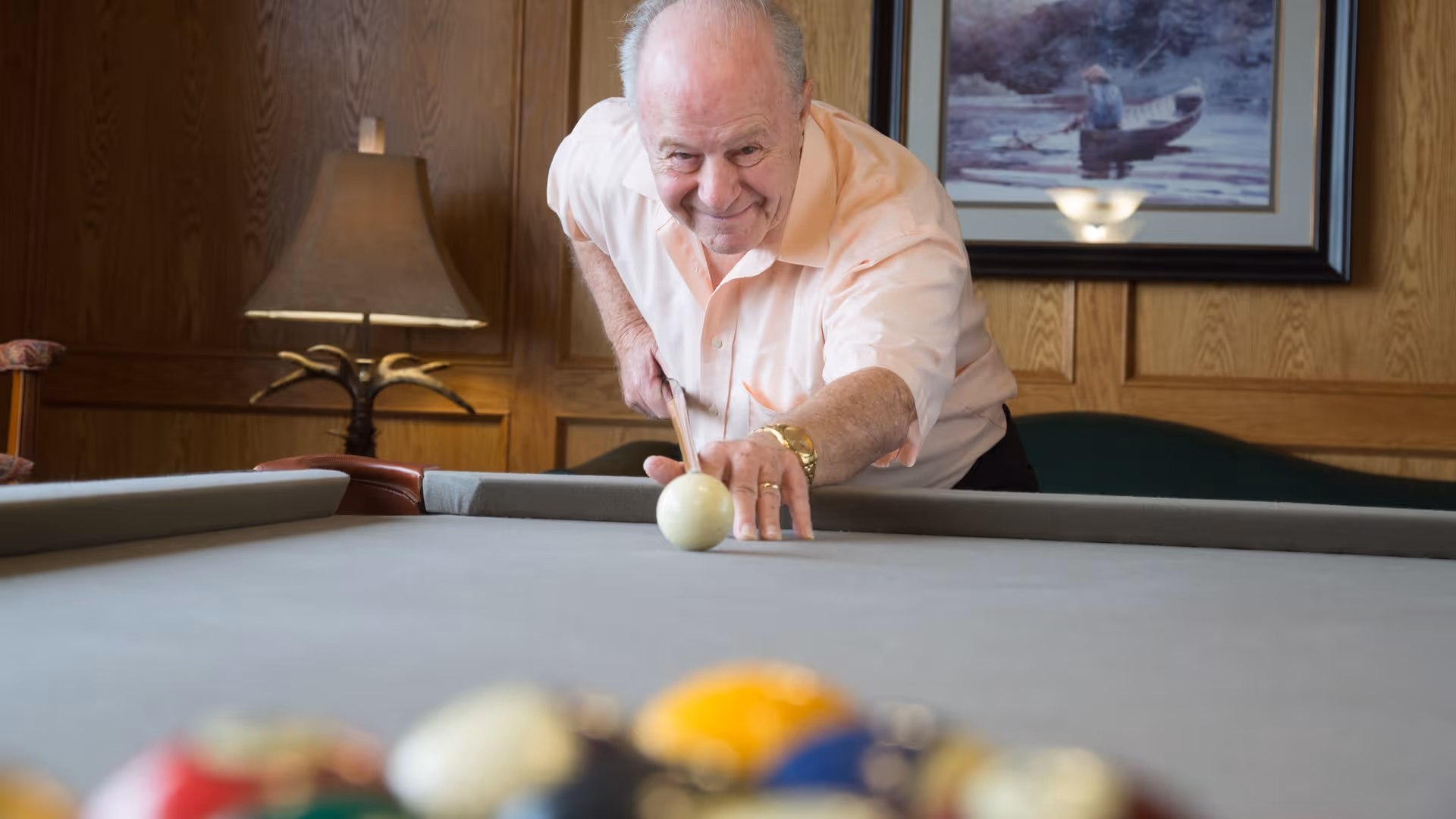 An elderly man playing pool indoors, aiming to strike the cue ball on a billiards table in a room with wood-paneled walls, a lamp, and a framed picture in the background.