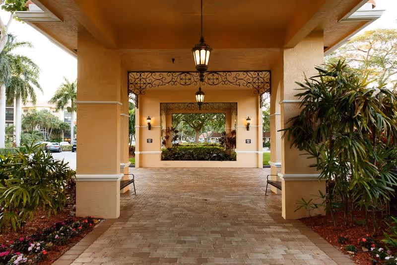Covered porte-cochere entrance to a beige building with hanging lanterns, columns, benches, and landscaping.