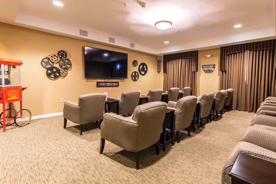 A cozy senior living facility media room with rows of cushioned armchairs facing a large flat-screen TV mounted on a beige wall. The room features carpeted flooring, decorative film reels on the wall, a popcorn machine on the left, and brown curtains covering the windows.