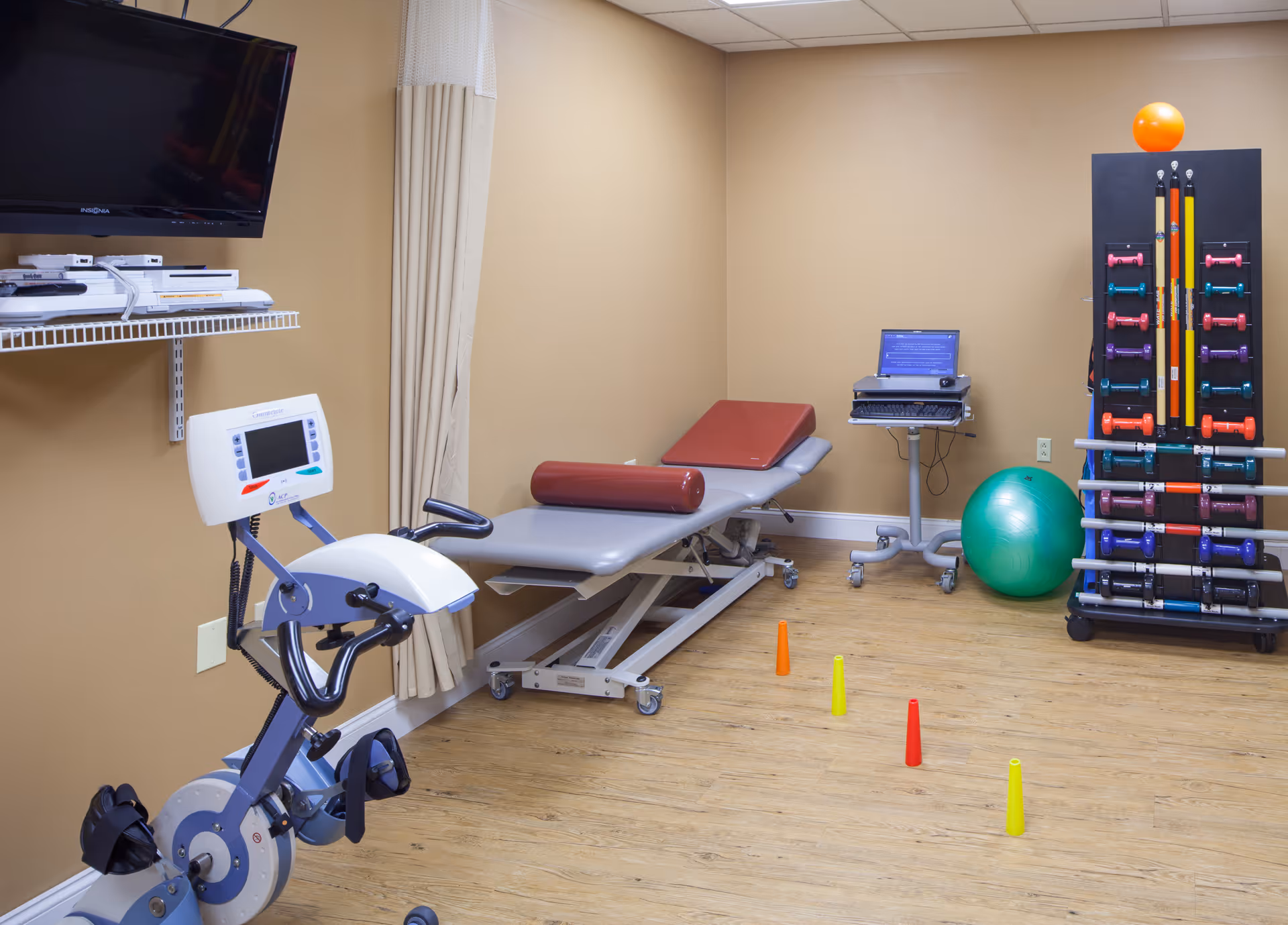 A physical therapy or exercise room with a stationary exercise bike, an adjustable therapy table with a cylindrical cushion, a laptop on a rolling stand, a rack holding various colorful dumbbells and exercise bars, a green exercise ball, and small colored cones placed on a wooden floor. The walls are beige and there is a TV mounted on the wall.