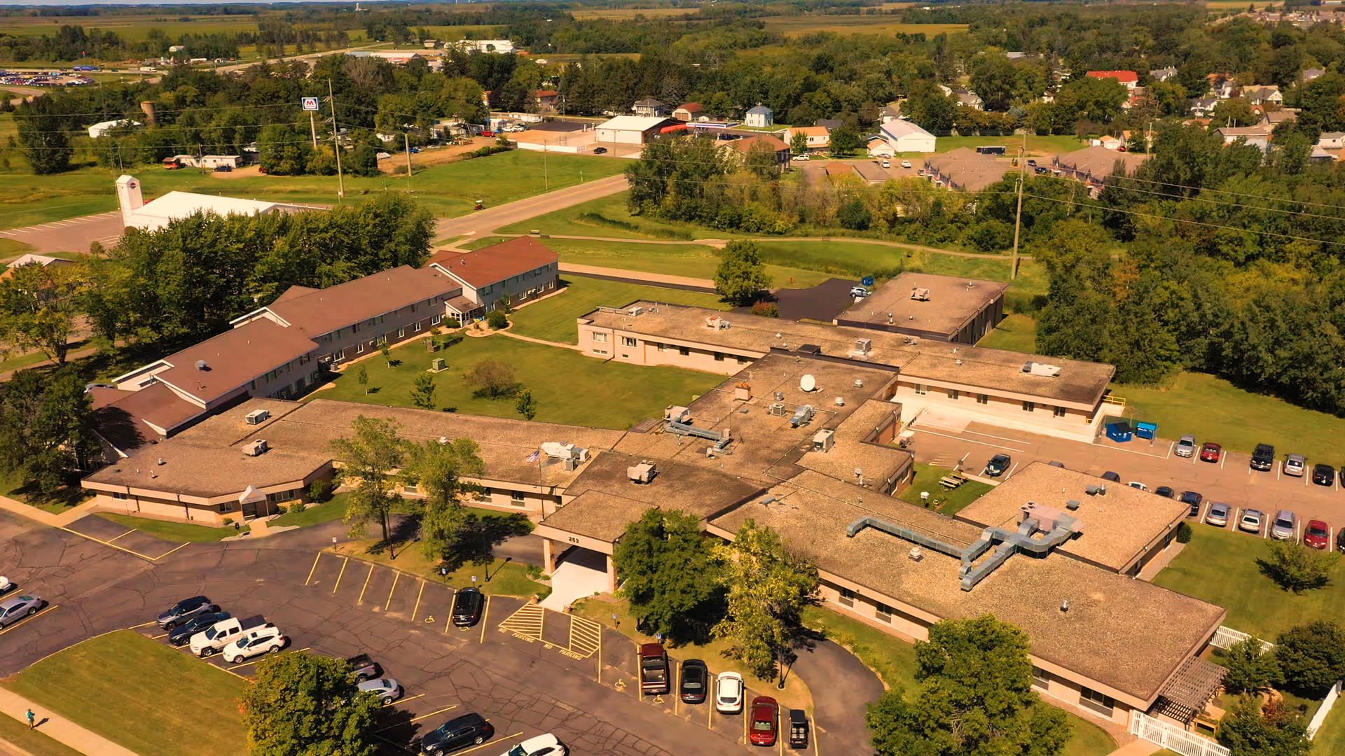 Aerial view of The Gardens at Foley showing a low-rise, multi-wing senior living complex with beige roofs, surrounding lawns, trees and parking lots.