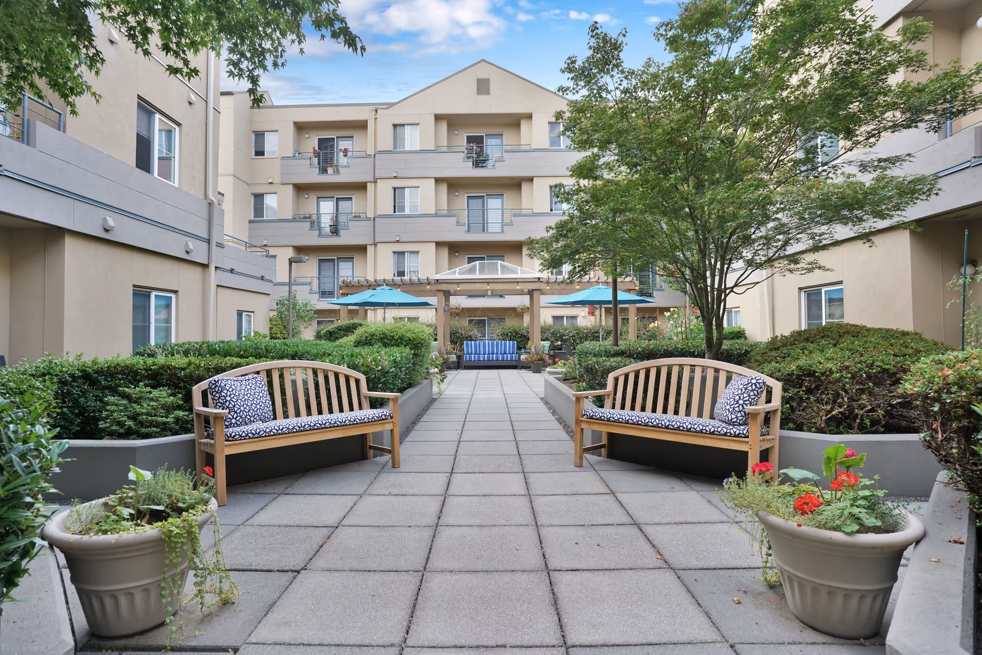 Courtyard of a multi-story assisted living building featuring a paved walkway, wooden benches with cushions, planters, umbrellas, and surrounding balconies.