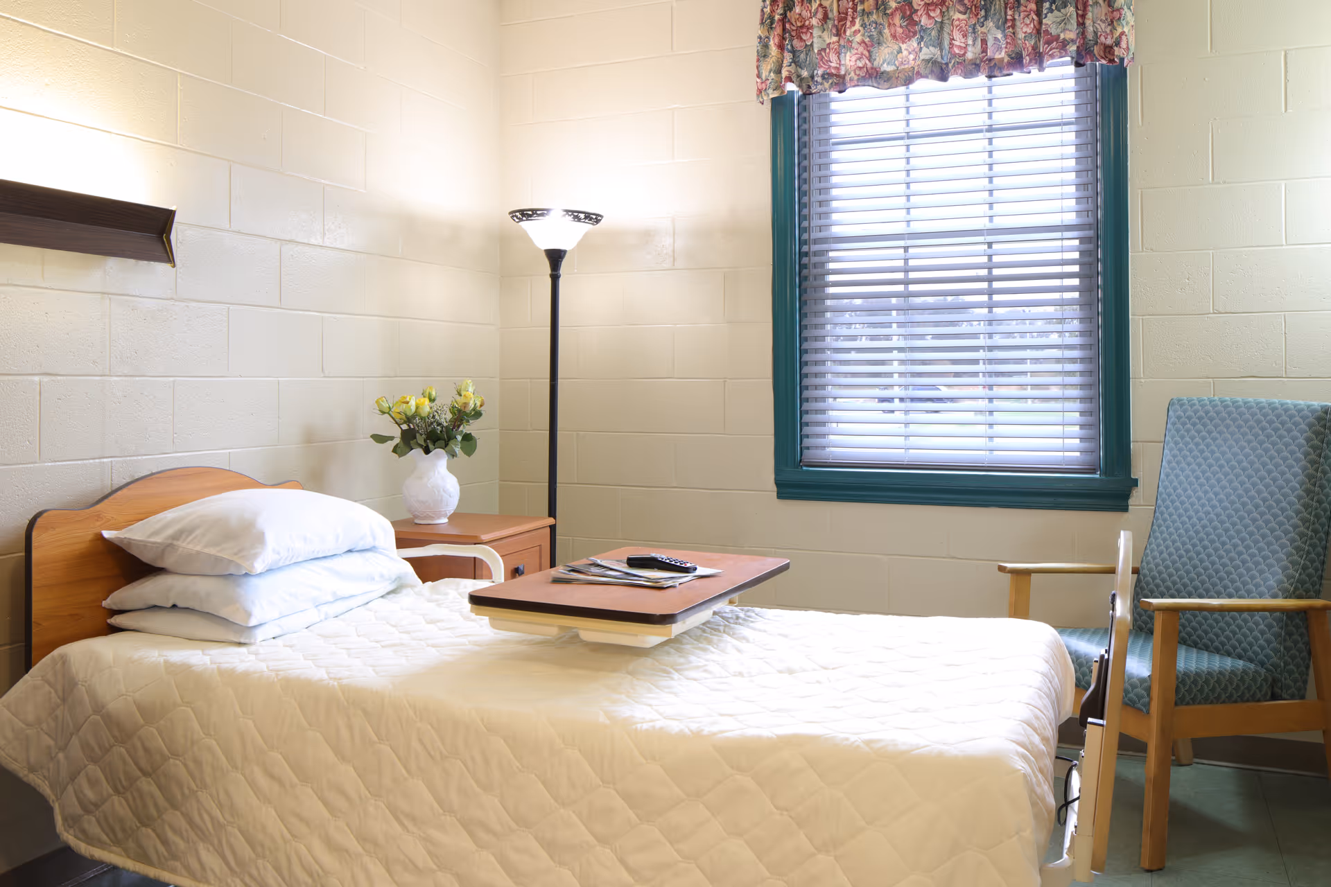A simple, clean bedroom in a senior living facility with a single bed covered in a white quilt, a wooden headboard, and three stacked pillows. Next to the bed is a wooden nightstand with a white vase holding yellow flowers. A tall floor lamp stands in the corner, and a window with blinds and a floral valance lets in natural light. A cushioned chair with wooden armrests is positioned near the window.