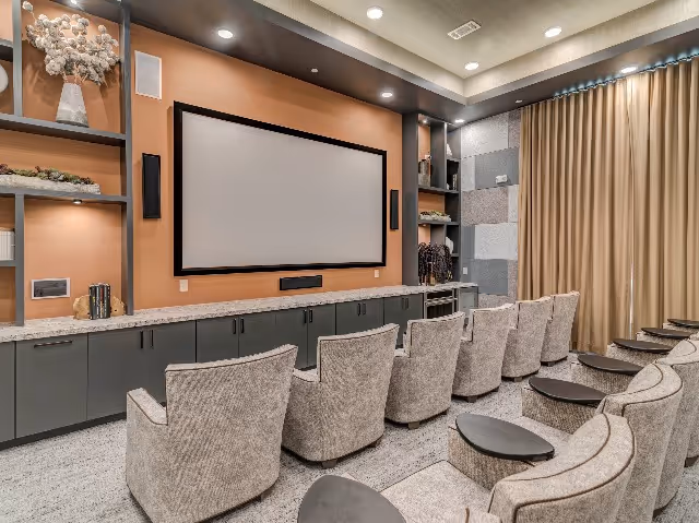 A modern media room with a large flat screen TV mounted on a wall above a long cabinet. There are two rows of comfortable upholstered chairs with small round tables in front of them. The room features built-in shelving with decorative items and soft lighting, and a large curtain covers one wall.
