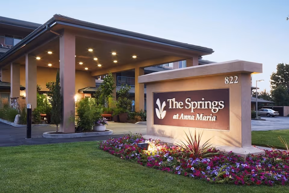 Entrance of The Springs at Anna Maria senior living facility with a large sign displaying the facility name, surrounded by well-maintained landscaping and flowers, and a covered drop-off area with lights on in the evening.