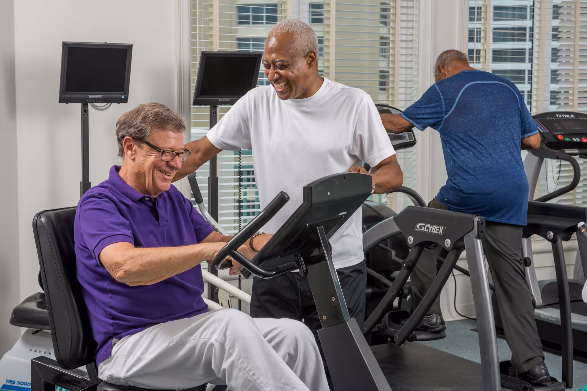 Three older men smiling and using cardio equipment in a bright fitness room.