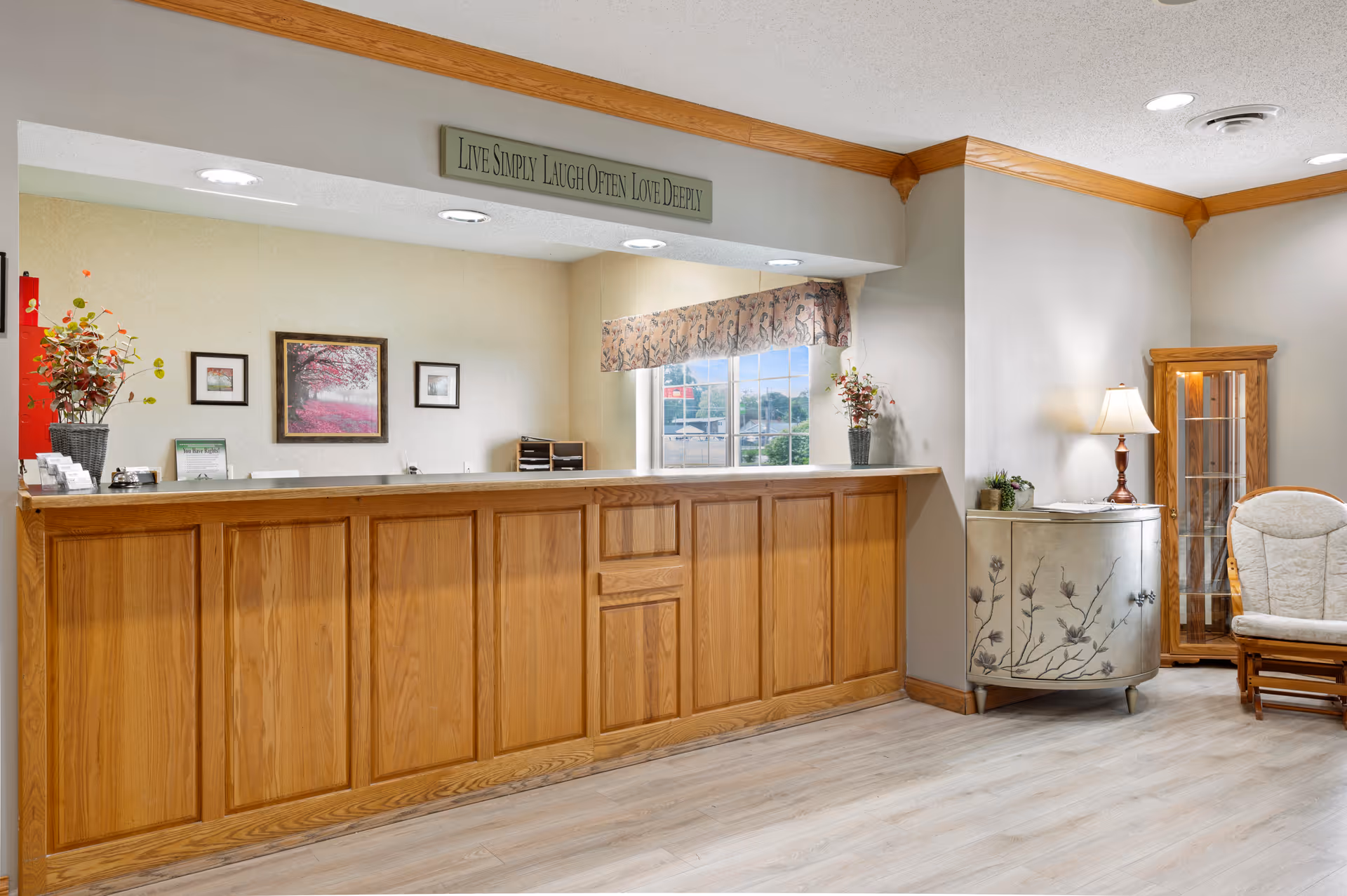 Wood-paneled reception desk and lobby area with a floral cabinet, lamp, seating, framed artwork, and a sign reading 'Live Simply Laugh Often Love Deeply'.