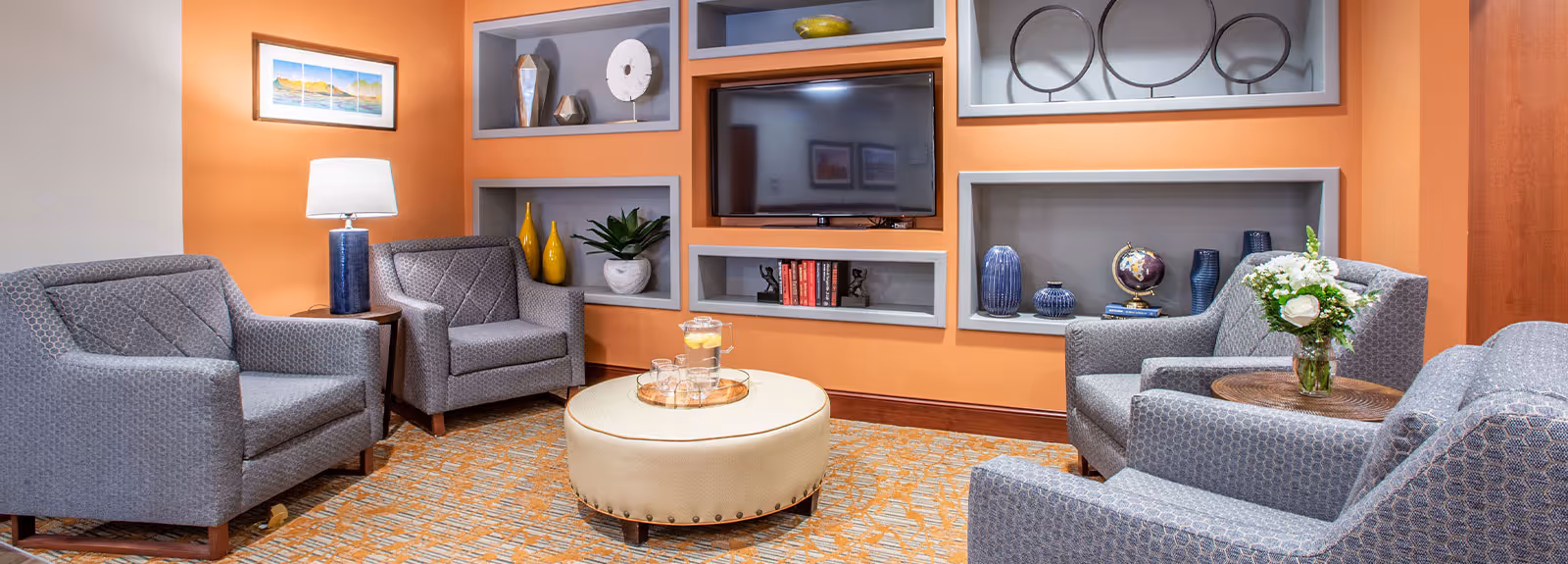 A cozy living room with four gray patterned armchairs arranged around a round beige ottoman with a tray holding a pitcher of water and glasses. The walls are painted orange with built-in shelves displaying decorative items such as vases, books, and a globe. A flat-screen TV is mounted on the wall above the shelves. There is a table lamp on a small side table between two chairs and a vase with white flowers on another side table.