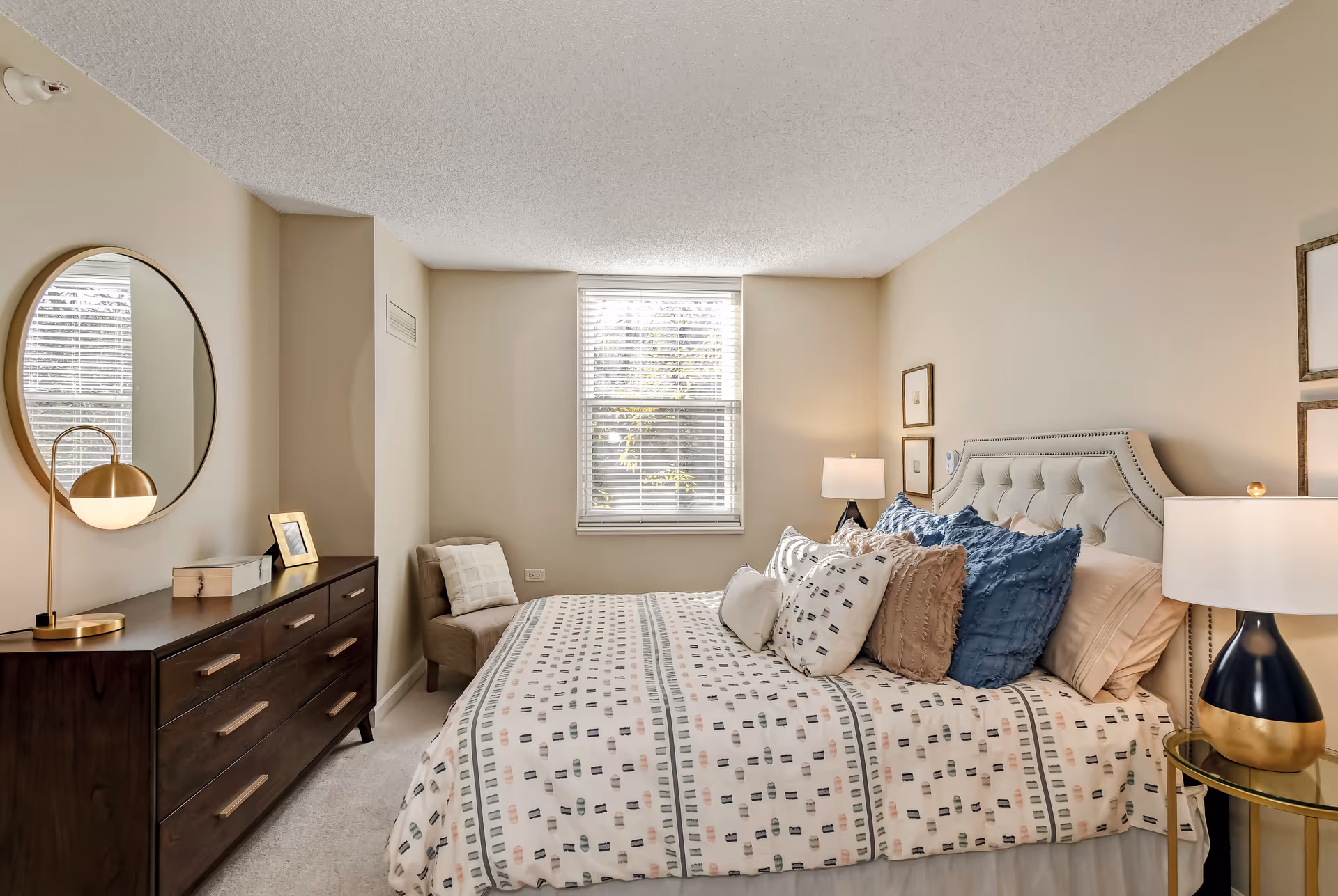 A cozy bedroom with a large bed adorned with multiple pillows and a patterned bedspread. There is a window with blinds letting in natural light, a cushioned chair in the corner, a dark wooden dresser with a round mirror above it, and two bedside lamps on either side of the bed.