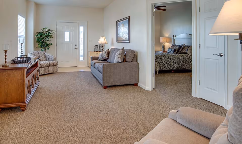 A cozy living room area with a gray sofa, an armchair, a wooden cabinet, a lamp, and a potted plant near the front door. The room has beige carpet and light-colored walls. An open doorway reveals a bedroom with a bed, nightstand, and lamp.