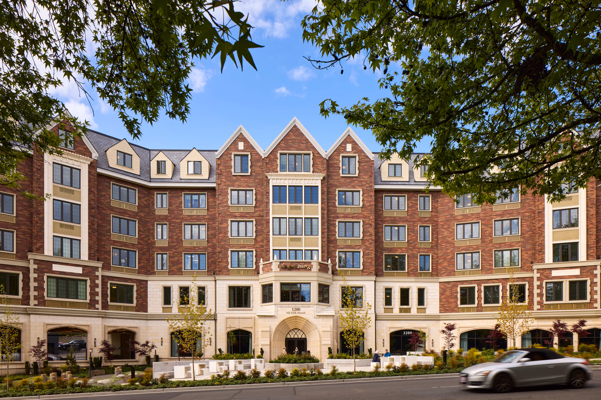 Front exterior view of Aegis Living Laurelhurst, a multi-story brick building with large windows and a central entrance. The building is framed by green tree branches and a blue sky with some clouds. There are small trees and landscaped bushes in front of the building, and a silver convertible car is driving on the road in the foreground.