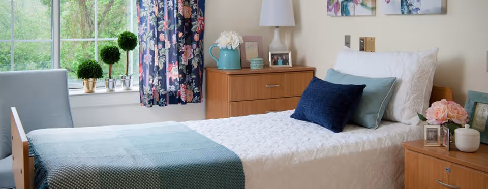 A cozy bedroom with a single bed made with white bedding and blue pillows. There is a wooden nightstand beside the bed with a vase of flowers, framed photos, and a small container. Another wooden dresser with a lamp, framed photo, and decorative items is next to the bed. A window with floral curtains and small potted plants on the windowsill lets in natural light.