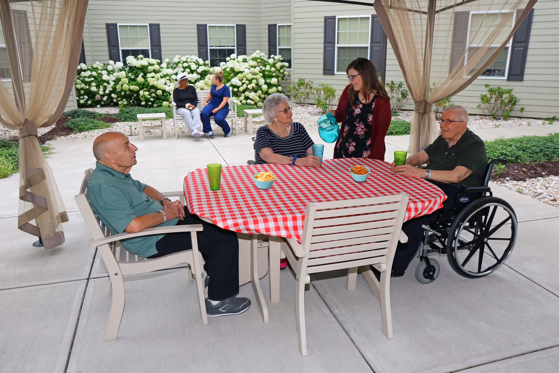 Three elderly people sitting around a table with a red and white checkered tablecloth outdoors under a canopy. A woman is pouring water into a glass for one of the elderly women. Two other women are sitting on a bench in the background near white flowering bushes and a building with light green siding and black shutters.