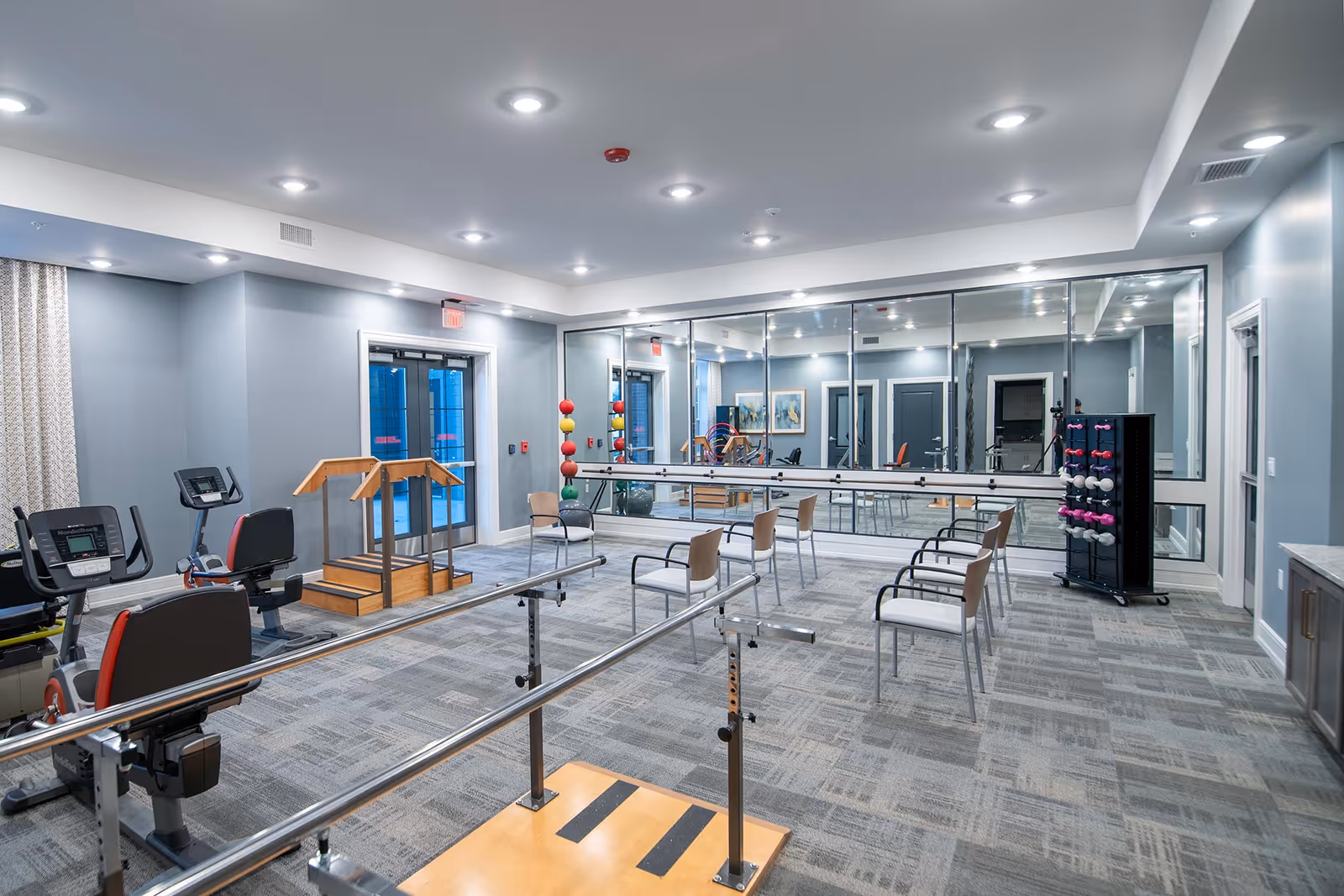A spacious exercise room in a senior living facility featuring parallel bars for walking support, several chairs arranged in a row, exercise bikes, a rack with colorful dumbbells, and a large wall mirror reflecting the room.