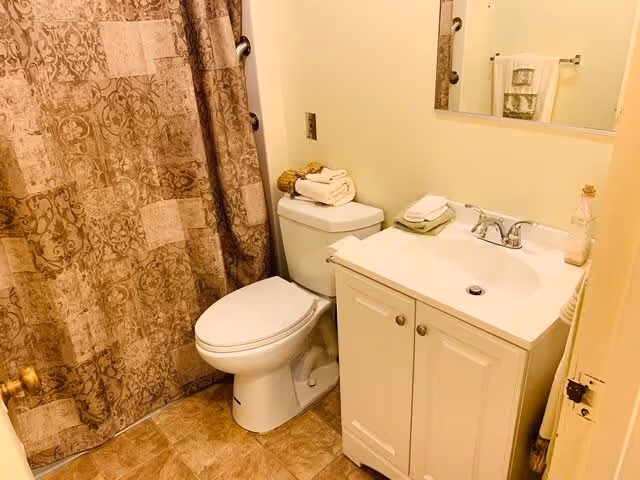 A small bathroom featuring a white toilet with folded towels on top, a white vanity with a sink and faucet, a mirror above the sink, and a shower with a patterned brown curtain. The floor is tiled in a light brown color.