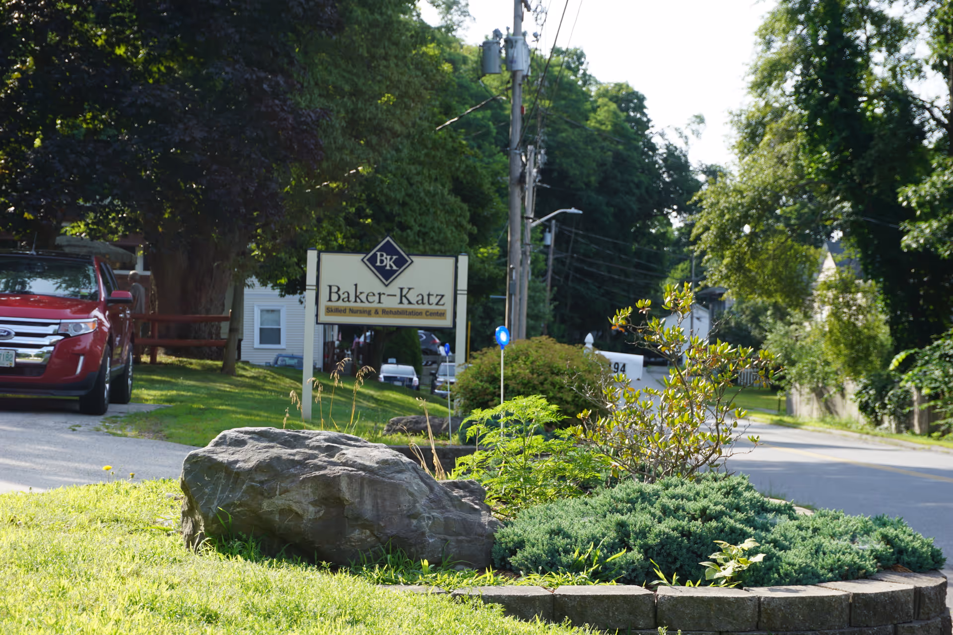Outdoor view of the entrance area to Baker-Katz Skilled Nursing & Rehabilitation Center, featuring a sign with the facility's name, a landscaped garden with rocks and bushes, a red car parked on the left, and a street lined with trees and houses in the background.