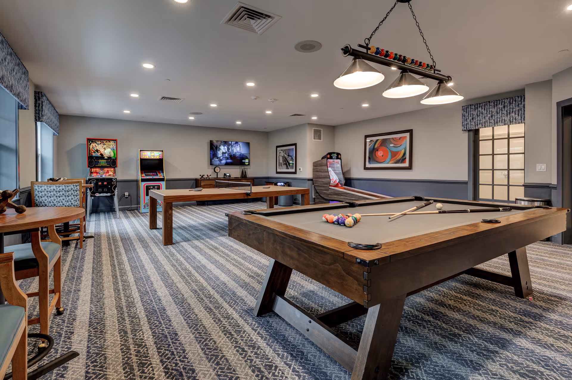 A recreational game room featuring a pool table with billiard balls and cues, a ping pong table, arcade game machines including Ms. Pac-Man and skee-ball, a wall-mounted TV, and seating with patterned chairs. The room has patterned carpet flooring, soft lighting, and framed artwork on the walls.