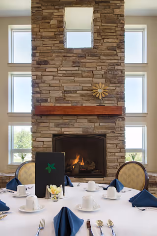 A dining table set with white tablecloth, white cups, plates, silverware, and navy blue napkins in front of a large stone fireplace with a wooden mantel and a decorative sun ornament. The fireplace is flanked by multiple windows letting in natural light.