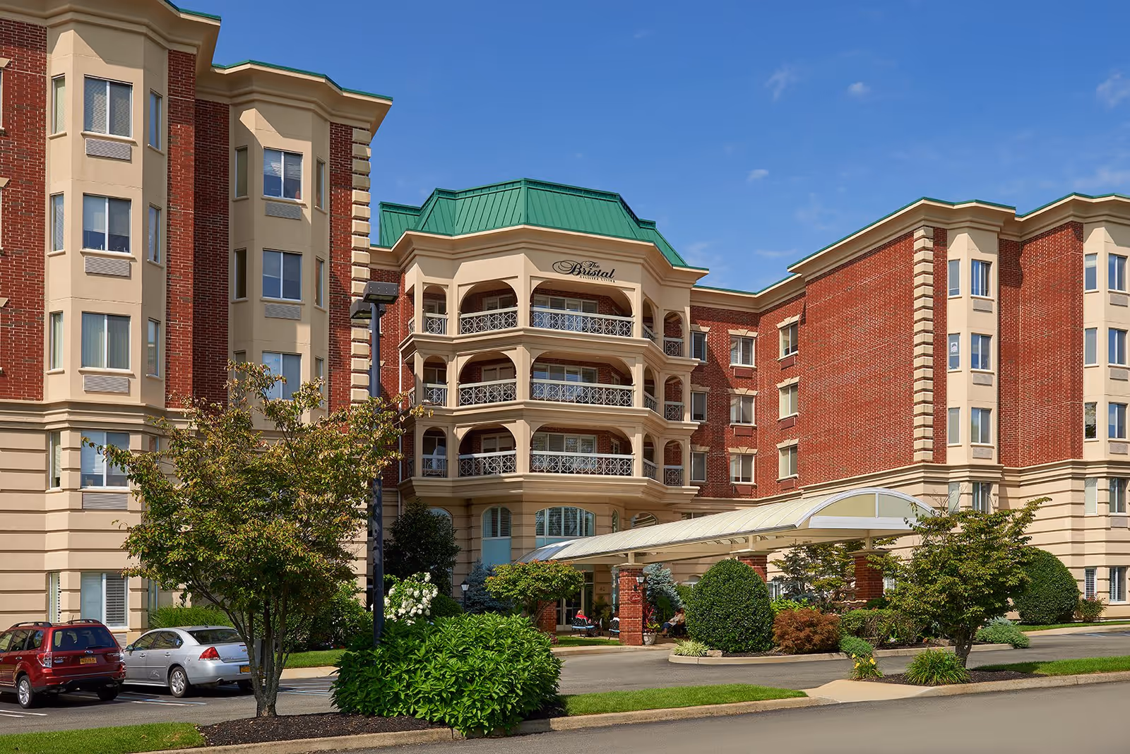 Exterior view of The Bristal Assisted Living at East Meadow building with red brick and beige facade, green roof accents, balconies, a covered entrance, landscaped bushes and trees, and parked cars under a clear blue sky.