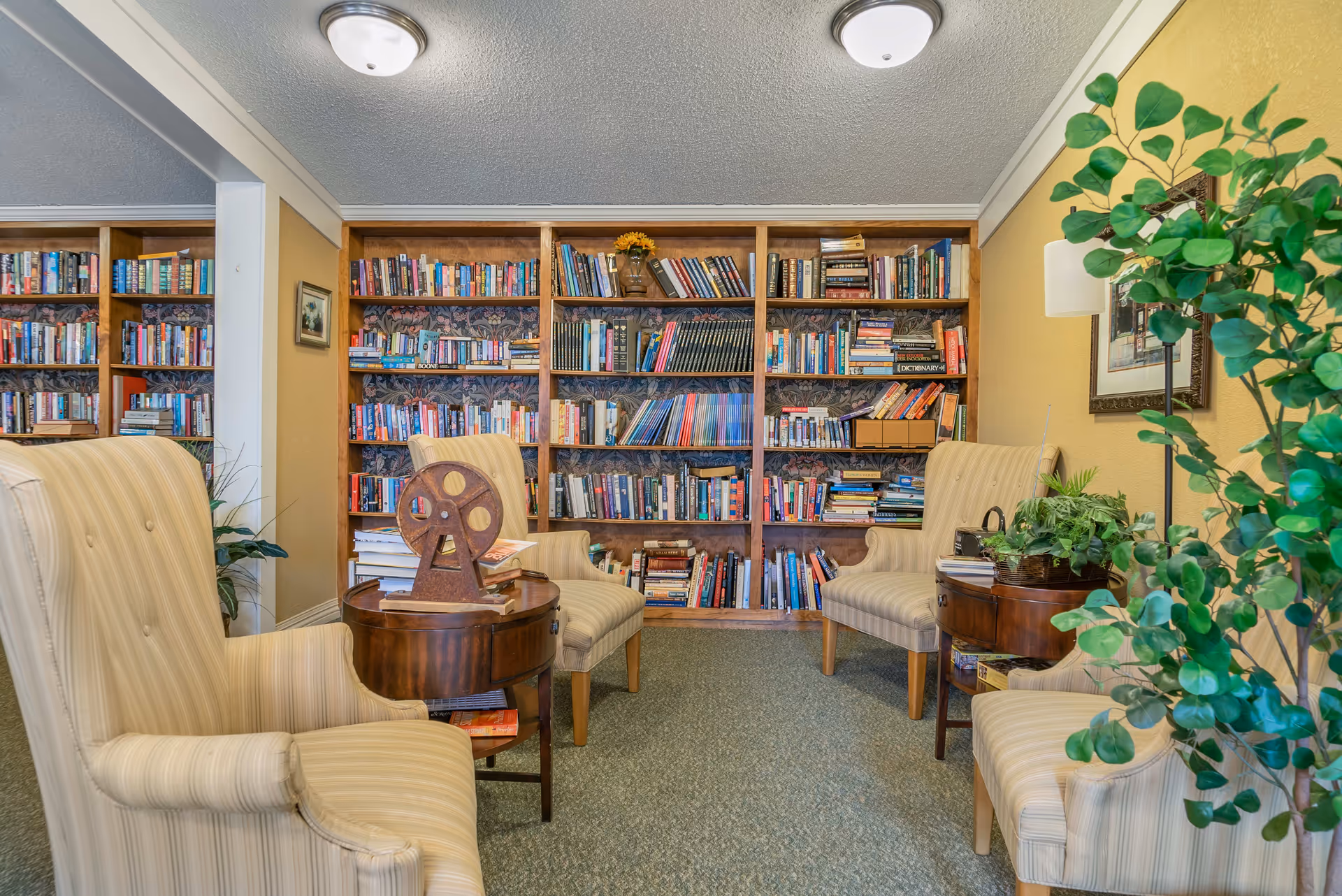 A cozy reading area with four beige striped armchairs arranged around two small wooden tables. Behind the chairs is a large wooden bookshelf filled with various books. There is a leafy green plant on the right side and a mirror on the left wall reflecting part of the bookshelf.