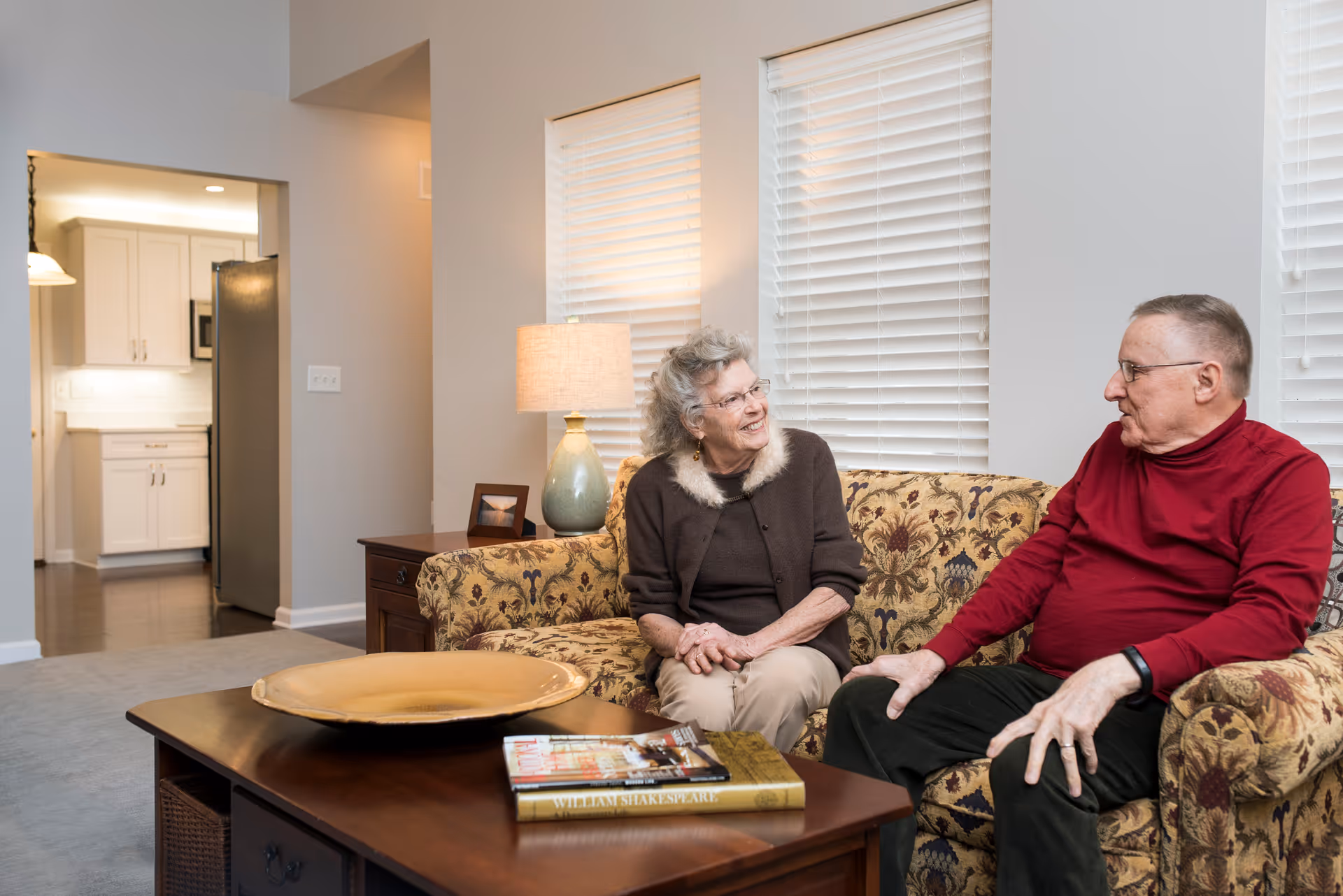 An elderly man and woman sit and talk on a patterned sofa in a well-lit living room with a coffee table and lamp.