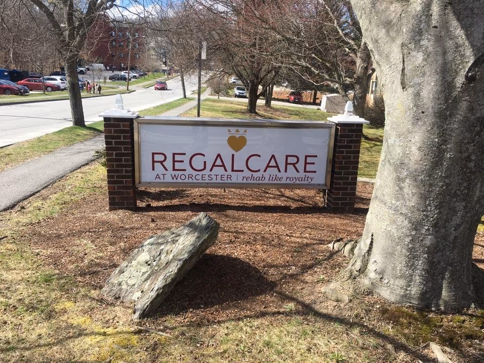Outdoor view of a sign for RegalCare at Worcester, placed between two brick pillars with a large tree and rock nearby. A street with parked cars and trees is visible in the background.
