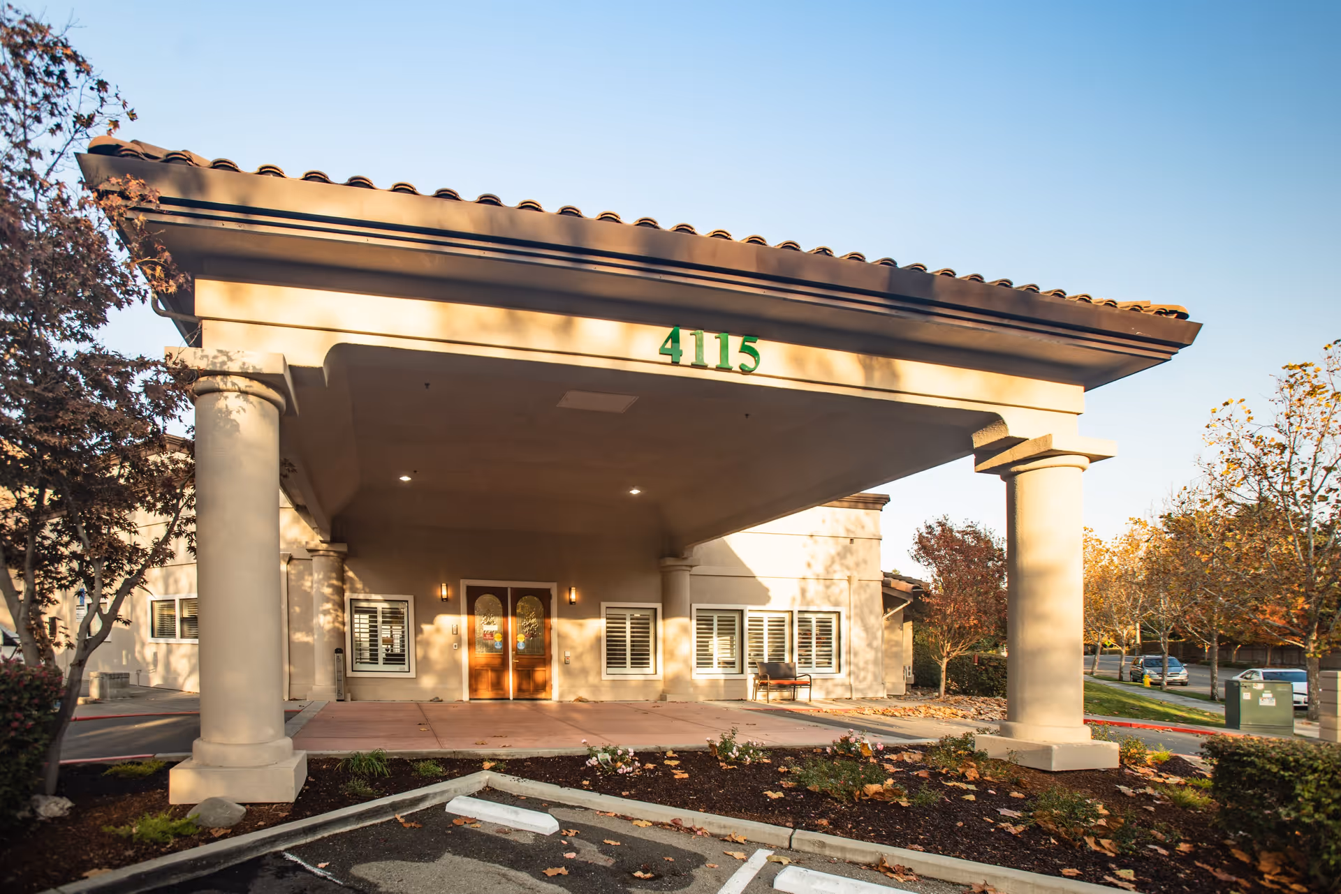 Front exterior view of Carefield Pleasanton Memory Care building entrance with a covered driveway supported by large columns, surrounded by trees and landscaping under a clear sky.