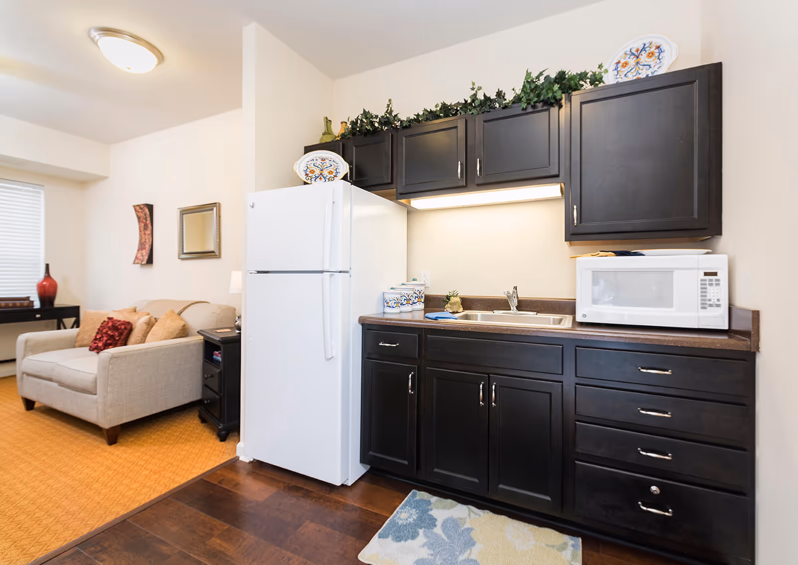A small kitchen area with dark wood cabinets, a white refrigerator, a white microwave on the countertop, and a stainless steel sink. Decorative plates and greenery are placed above the cabinets. Adjacent to the kitchen is a living area with a beige loveseat, red and beige pillows, a side table with a lamp, and wall decorations.