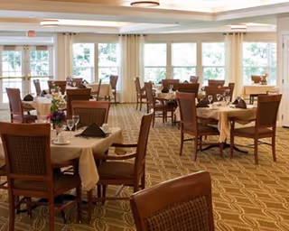 A spacious dining room with multiple round tables covered with beige tablecloths, each set with glassware, napkins, and floral centerpieces. The room has large windows allowing natural light to fill the space, and wooden chairs with cushioned seats surround the tables.