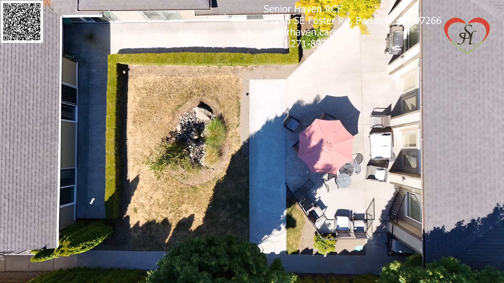Aerial view of an outdoor courtyard area at Senior Haven RCF featuring a central patch of dry grass with a small rock and plant arrangement, surrounded by concrete walkways and green hedges. There is a patio area with outdoor furniture including chairs, tables, and a red umbrella providing shade. The surrounding buildings have multiple windows and doors facing the courtyard.