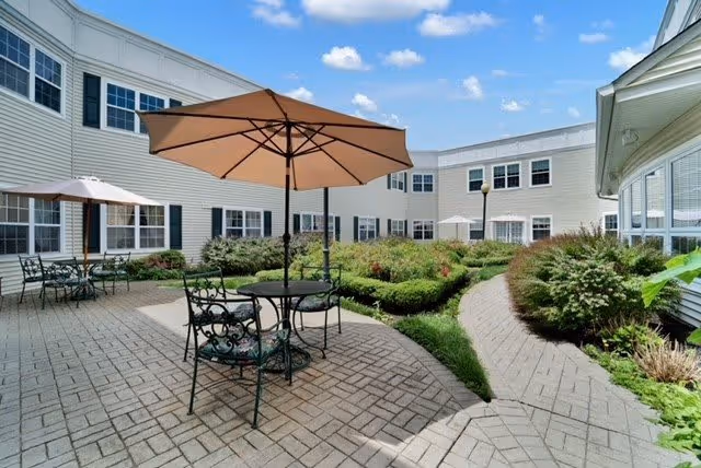 Outdoor courtyard area at a senior living facility with paved walkways, green shrubs, and patio tables with umbrellas under a blue sky with scattered clouds.