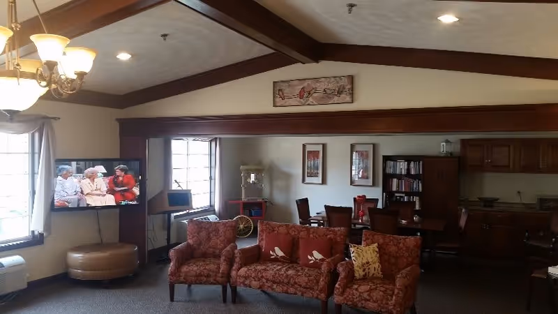 A cozy living room area in an assisted living facility with patterned armchairs and a sofa, a flat-screen TV mounted on the wall showing a program with three elderly people, a dining table with chairs, a popcorn machine, bookshelves, and wooden cabinetry. The room has exposed wooden beams on the ceiling and large windows letting in natural light.